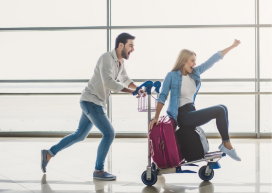 Man pushing woman on luggage trolley through airport looking excited