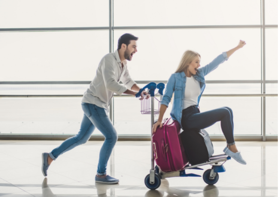 Man pushing woman on luggage trolley through airport looking excited Man pushing woman on luggage trolley through airport looking excited